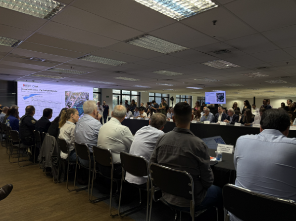 A imagem mostra uma sala de reuniões ampla durante uma apresentação formal. No primeiro plano, um grupo de pessoas está sentado de costas para a câmera, ocupando cadeiras pretas ao longo de uma mesa comprida e escura. Os participantes vestem roupas em tons neutros, como camisas sociais azuis, brancas e cinzas. Ao fundo, do lado esquerdo, uma grande tela de projeção exibe um slide com o título "Estudo de caso - Pq. Independência", acompanhado de um gráfico e um mapa técnico. À direita, há um monitor menor suspenso com informações complementares. O ambiente é iluminado por luminárias embutidas em um teto de placas brancas e possui janelas ao fundo que revelam a claridade do dia. Sobre a mesa, em frente aos participantes, nota-se uma placa de identificação com o texto "DESENVOLVIMENTO ECONÔMICO E TRABALHO".