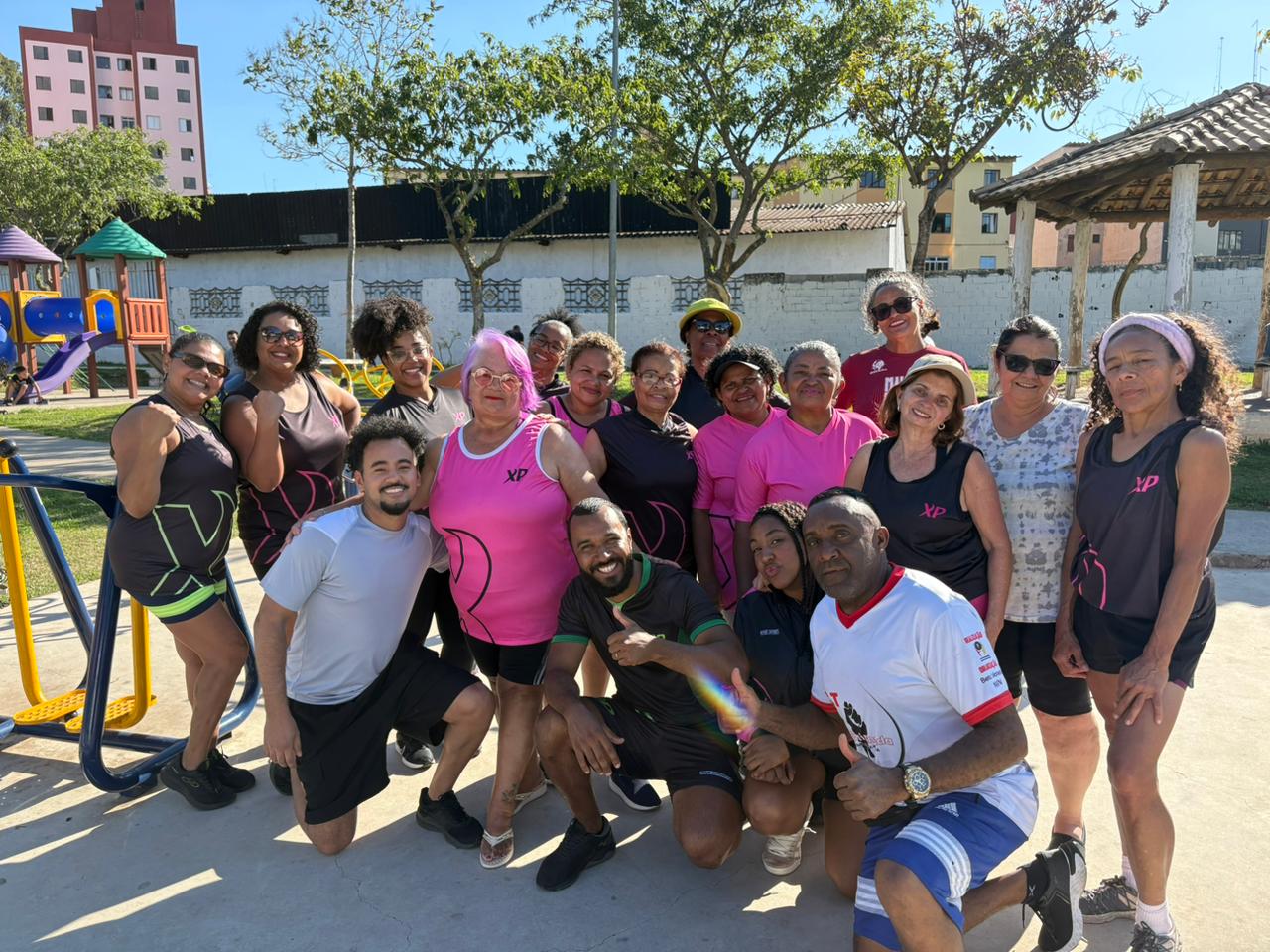 A fotografia em plano médio captura um grupo heterogêneo de aproximadamente dezessete pessoas posando juntas, em sua maioria mulheres, em uma área externa bem iluminada, possivelmente uma praça ou parque. O grupo está centralizado e sorridente, com a maioria das mulheres vestindo roupas de ginástica em tons de preto, cinza e rosa choque (algumas com a sigla "XP" visível).  Quatro homens estão misturados no grupo: dois agachados na frente e dois em pé mais atrás, com um dos homens em pé usando uma camiseta esportiva branca e azul. O grupo está disposto em três ou quatro fileiras informais, olhando diretamente para a câmera, transmitindo uma sensação de comunidade e atividade física.  Ao fundo, a área é definida por piso de cimento claro no primeiro plano. Imediatamente atrás do grupo, nota-se uma área de lazer, com brinquedos coloridos de um parquinho infantil à esquerda e um quiosque de madeira com teto branco à direita. Árvores frondosas sombreiam a parte superior da cena, e edifícios residenciais e muros baixos compõem o plano de fundo. O dia está ensolarado.
