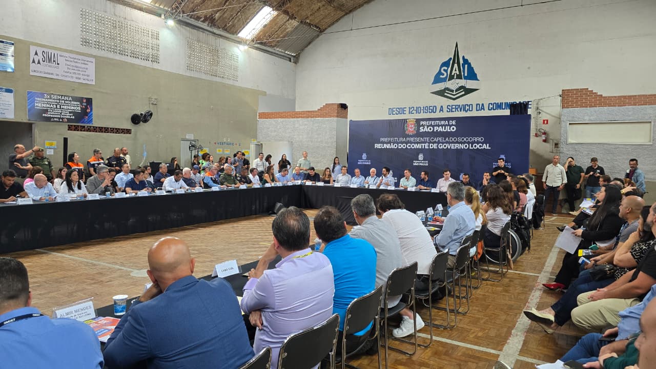 Close ainda mais detalhado de um trecho da mesa central, mostrando autoridades em discussão, uma delas falando ao microfone. Ao fundo, várias pessoas acompanham a reunião de pé, algumas fazendo registros com celular, reforçando o clima de evento técnico e público.