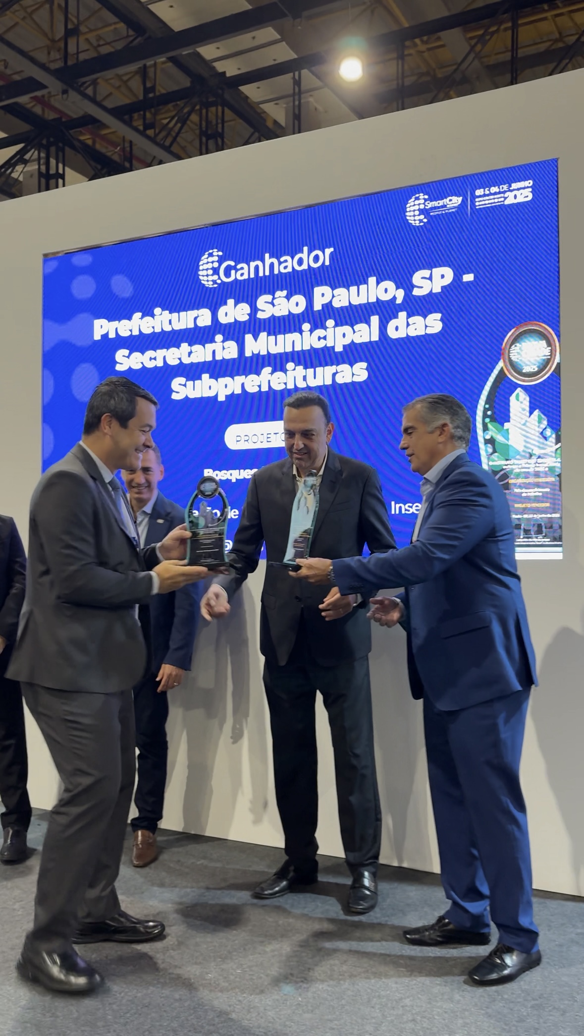 A imagem mostra três homens adultos em um palco ou evento formal, com um grande painel de LED azul ao fundo. O painel exibe o texto "Ganhe a Prefeitura de São Paulo, SP - Secretaria Municipal das Subprefeituras" em destaque, indicando uma premiação ou reconhecimento.  No centro da foto, um homem de terno escuro (ao meio) está recebendo um troféu de vidro ou acrílico de formato orgânico, que parece ser um prêmio, entregue por um segundo homem (à esquerda) também de terno escuro, que está de perfil. Ambos se olham enquanto um terceiro homem (à direita), de terno azul escuro, também se aproxima e parece estar envolvido na entrega ou recebimento do prêmio.  Os três homens parecem profissionais, com sorrisos sutis, indicando um momento de celebração ou cerimônia. O ambiente ao redor é formal e bem iluminado. Há outros homens desfocados ao fundo, sugerindo a presença de mais participantes ou público.